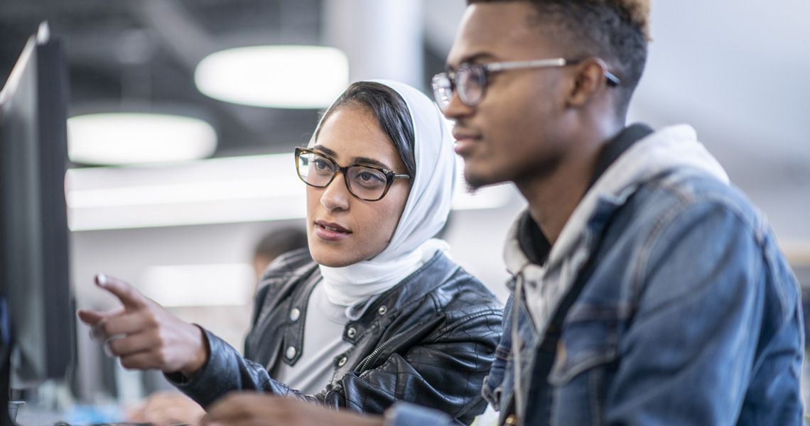 Two diverse students collaborate at a computer in a modern research setting, analyzing data. Their discussion reflects the interdisciplinary teamwork essential in a Clinical and Translational Research Program, where scientific discoveries are translated into medical advancements.
