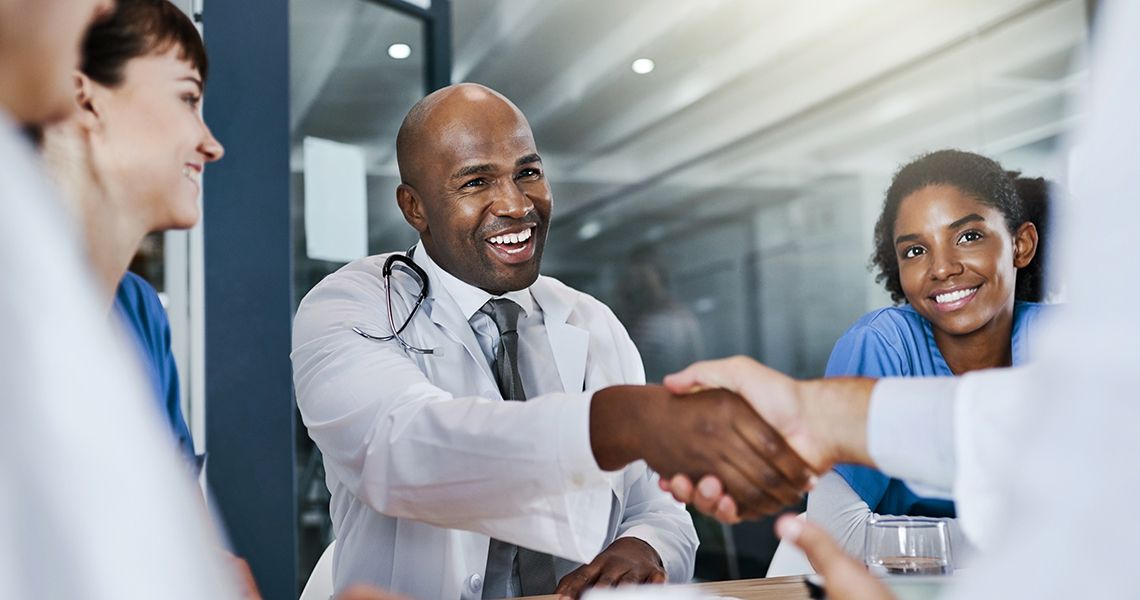 doctor with a stethoscope around his neck smiling and shaking hands with someone across a table