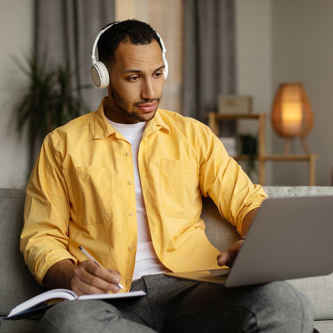 A person wearing headphones using their laptop and writing in a notebook