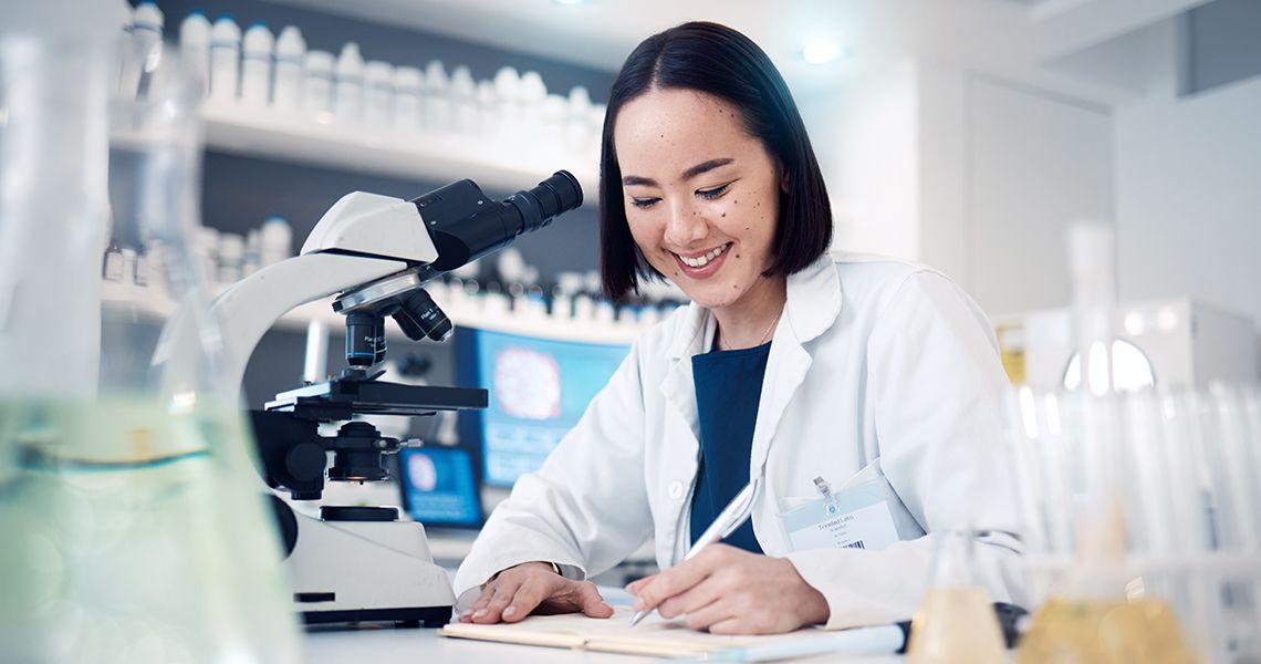 A female researcher is writing while seated next to a microscope