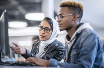 Two diverse students collaborate at a computer in a modern research setting, analyzing data. Their discussion reflects the interdisciplinary teamwork essential in a Clinical and Translational Research Program, where scientific discoveries are translated into medical advancements.