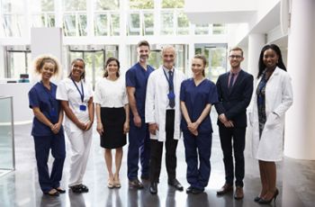 A diverse group of healthcare professionals and administrators, including nurses, doctors, and business staff, standing together and smiling in a bright, modern medical facility—illustrating teamwork and leadership in clinical operations and health care management.